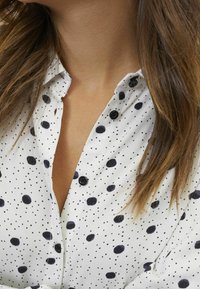 Weiße Bluse mit schwarzen Polka-Dots, geknöpfter Kragen, aus einem weichen Material mit leichtem Glanz. Nahaufnahme des Ausschnitts und des Stoffs.