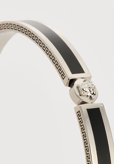 Ασημένιο βραχιόλι με μαύρες επενδύσεις, σχέδιο ελληνικού κλειδιού κατά μήκος των άκρων και λεπτομέρεια κεφαλής λιονταριού στην αγκράφα, πάνω σε λευκό φόντο.