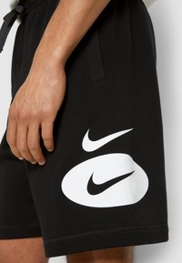 Shorts en coton noirs avec un grand logo Nike blanc sur le côté, dotés d'une taille élastique et de poches latérales. Vue rapprochée de la main.