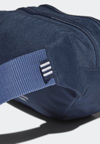 Námorně modrý látkový pytlík s texturovaným povrchem, vybavený pevným popruhem a výrazným bílým pruhem. Zipové zavírání v horní části.