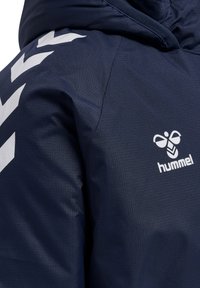 Chaqueta azul marino con tejido texturizado, con patrones angulares blancos y un logo prominente en el pecho. Diseño con capucha.