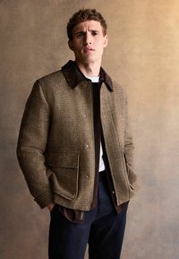 Brown houndstooth jacket with a dark brown suede collar, snap closure, two front pockets, and layered over a dark shirt and white T-shirt.