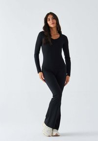 Schwarzer Bodysuit mit langen Ärmeln und U-Ausschnitt, kombiniert mit eng anliegenden, ausgestellten Hosen aus glattem Stoff. Dazu helle Sneaker getragen.