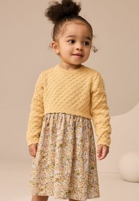 Pull en maille jaune avec un motif texturé, associé à une robe à motif floral présentant des fleurs roses, bleues et vertes sur un fond clair.