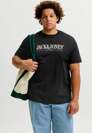 Ung mand med krøllet hår iført en sort Jack & Jones t-shirt og lyseblå jeans, der holder en grøn og hvid mulepose over skulderen.