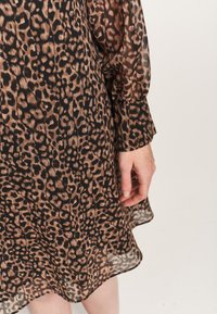 Vestido de gasa con estampado de leopardo y mangas largas y transparentes. La tela presenta una mezcla de marrones y negros, destacando una textura fluida y ligera.