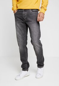 Henkilö, jolla on päällään haalistuneet mustat farkut ja valkoiset tennarit, kädet taskuissa, keltaisessa pitkähihassa olevaa paitaa yksinkertaista taustaa vasten.