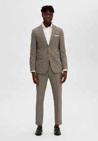 Un costume gris clair à carreaux avec une coupe ajustée, une veste à un bouton, des revers crantés, deux poches à l'avant et un pantalon assorti. Chaussures noires.