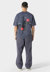 Granatowa bawełniana koszulka z grafiką żurawia w czarnych i czerwonych okręgach na plecach, połączona z luźnymi szarymi spodniami dresowymi.