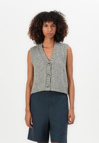 Jonge vrouw met krullend haar in een grijze mouwloze gebreide vest met knopen en donkere knielange shorts, die recht vooruit kijkt.