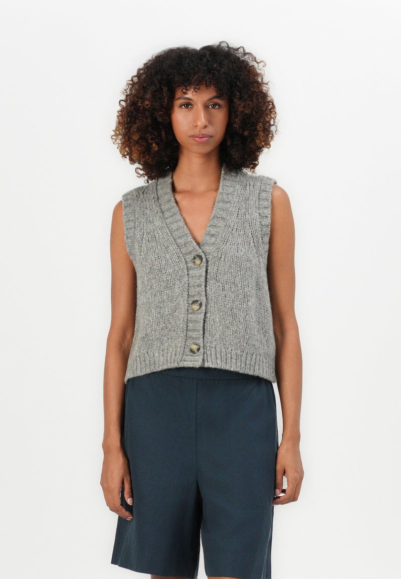Jonge vrouw met krullend haar in een grijze mouwloze gebreide vest met knopen en donkere knielange shorts, die recht vooruit kijkt.