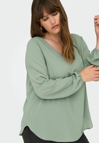Nano zelena bluza z V-izrezom, dolgimi rokavi s nabranimi manšetami in lebdečo silhueto. Mehak material z rahlo ukrivljenim robom.