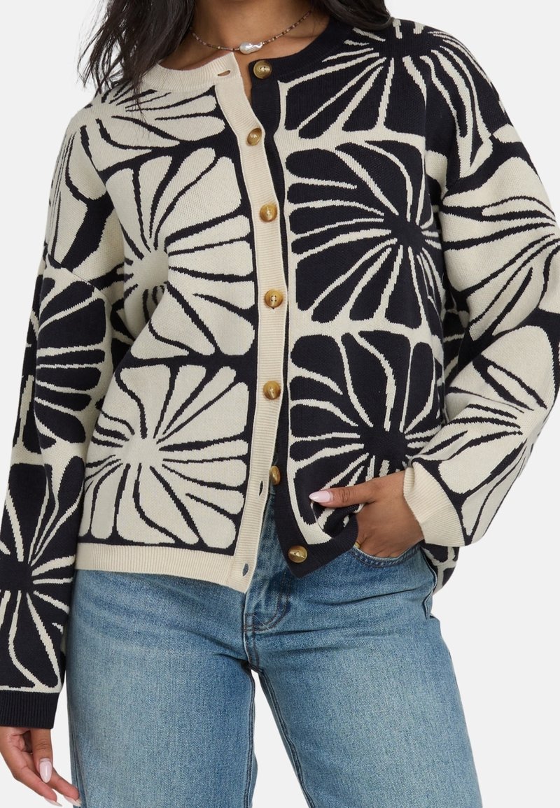 Femme portant un gilet boutonné noir et beige avec de grands motifs floraux, associé à un jean bleu clair et un collier de perles.