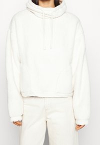 Biała, krótką bluza z kapturem, wykonana z miękkiego, puszystego materiału, z wysoką szyją, regulowanymi sznureczkami i szerokimi rękawami. Szczegóły przeszycia na dolnym brzegu.