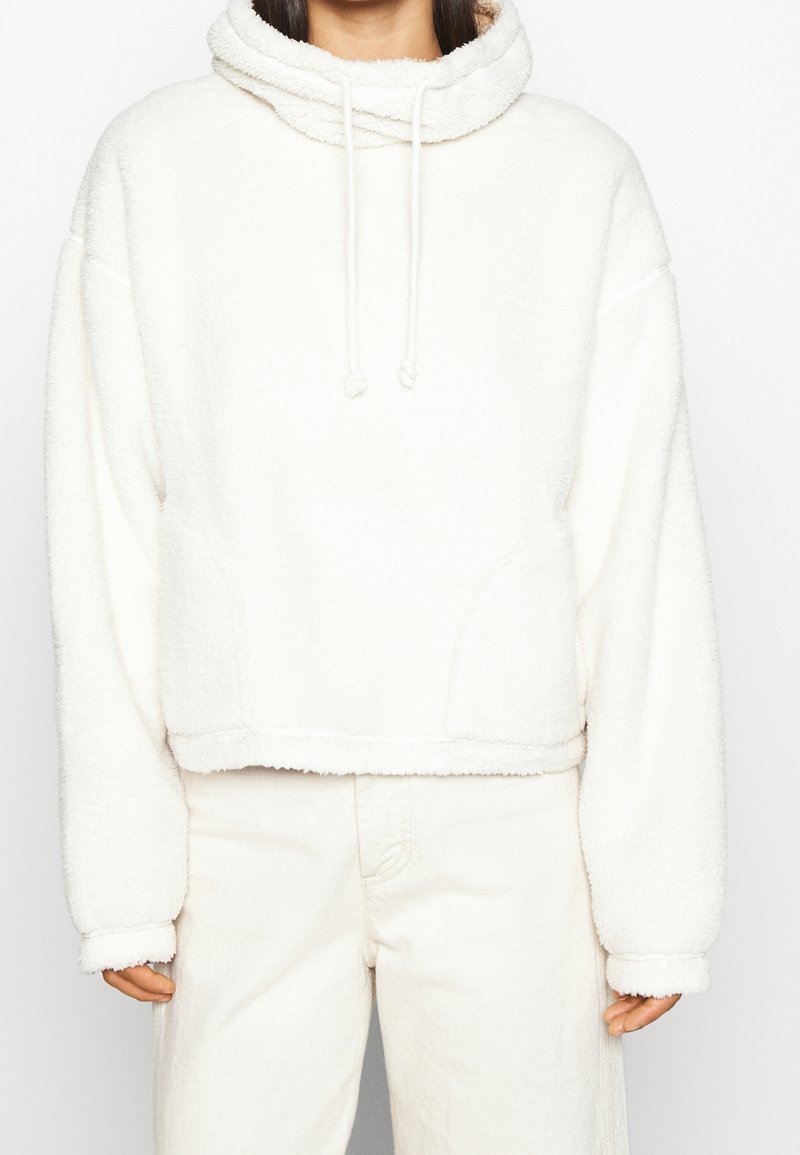 Biała, krótką bluza z kapturem, wykonana z miękkiego, puszystego materiału, z wysoką szyją, regulowanymi sznureczkami i szerokimi rękawami. Szczegóły przeszycia na dolnym brzegu.
