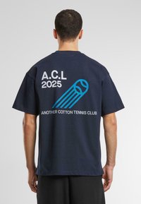 Tamsiai mėlynas medvilninis marškinėliai trumpomis rankovėmis, su balta ir mėlyna tekstu: "A.C.L 2025" ir "ANOTHER COTTON TENNIS CLUB", taip pat teniso kamuoliuko grafika.