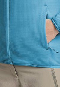 Veste bleu clair texturée avec une fermeture éclair à l'avant et une poche latérale. Cordon de serrage réglable à l'ourlet, assortie à un pantalon beige.