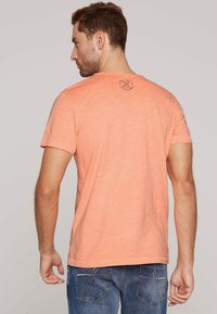 Koszulka z krótkim rękawem, okrągłym dekoltem, w jasnym koralowym kolorze z teksturowanym wykończeniem. Posiada okrągłą grafikę na górnej części pleców.