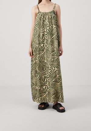 Persoon die een mouwloze, lange beige jurk draagt met donker groene grote bladpatronen en zwarte platformsandalen op een lichte achtergrond.