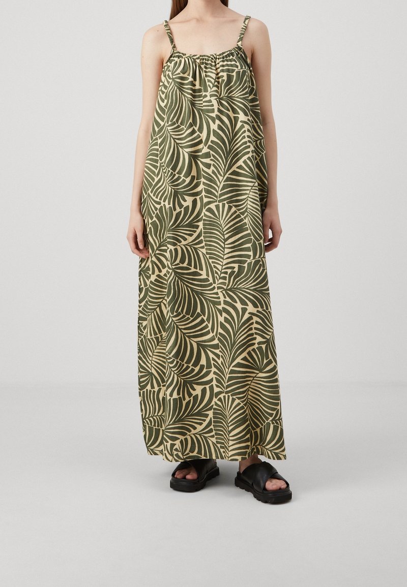 Personne portant une robe longue beige sans manches avec de grands motifs de feuilles vert foncé et des sandales noires à plateforme sur un fond clair.