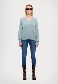 Pullover bleu deschis, cu decolteu în V, dintr-o textură moale, asortat cu blugi albaștri mulati și cizme lucioase maro până la genunchi. Ochelari de soare rectangulari negri purtați.