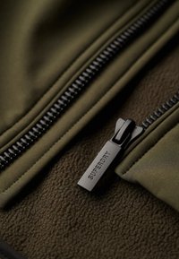 Tkanina polarowa w oliwkowym kolorze o gładkiej fakturze, z czarnym suwakiem oznaczonym napisem "SUPERDRY" oraz detalami ściegów.