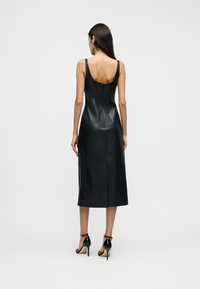 Μαύρο δερμάτινο midi φόρεμα με εφαρμοστή σιλουέτα, φαρδιές τιράντες, κλείσιμο με φερμουάρ στην πλάτη και λεία υφή. Φορεμένο με μαύρα πέδιλα με λουράκια.