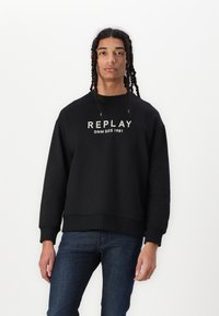 Černá mikina s nápisem "REPLAY" a "DNM GDS 1981" vytištěným bílou barvou na prsou. Hribované manžety a lem, volný střih. Nosit s tmavými džíny.