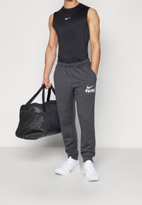 Black sleeveless top and gray sweatpants with a white Nike logo. Man holds a black duffel bag and wears white athletic shoes.