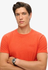 Ung mand med kort brunt hår iført orange T-shirt og sort armbåndsur, arme krydset, neutral ansigtsudtryk, ensfarvet hvid baggrund.