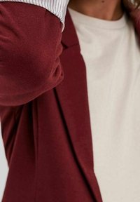 Burgundi bleiseri, jolla on sileä rakenne ja jämäkkä muotoilu, yhdistettynä kermaiseen t-paitaan, jossa on pyöreä pääntie ja näkyvät raidalliset hihansuut.