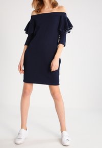 Granatowa sukienka z odkrytymi ramionami, z falbanowymi rękawami, dopasowany fason, długość do kolan i miękka tekstura, zestawiona z białymi butami wsuwanymi.