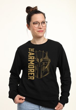 Kvinde med briller iført en sort sweatshirt med et gyldent Mandalorian-hjelm og teksten "The Armorer."