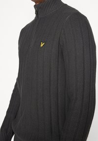 Dunkelgrauer strickpullover mit Reißverschluss am Hals, geripptem Muster und einem gestickten gelben Logo auf der linken Brust.