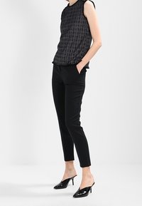 Černý vzorovaný bezrukávový top s postranním drapováním, doplněný úzkými černými kalhotami a lakovanými černými muly. Hladká tkanina a přiléhavý střih.