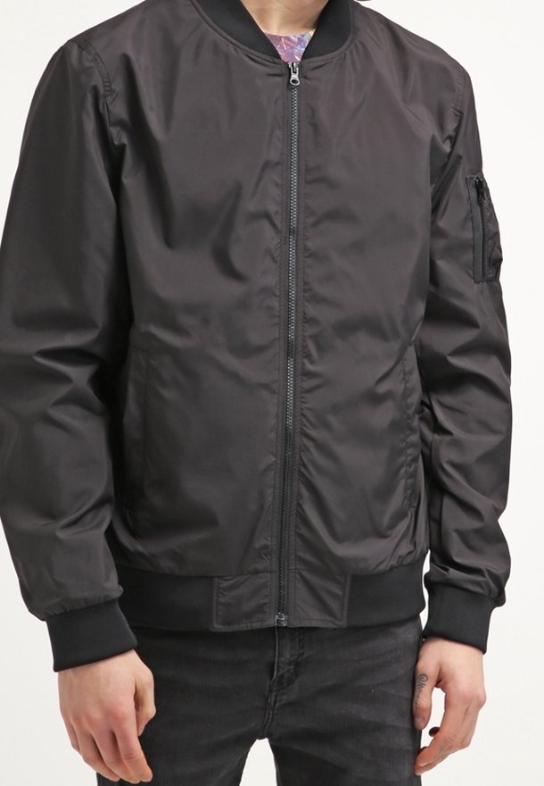 Person som bär en svart zip-up bomberjacka med ribbade ärmslut och nederkant, tillsammans med svarta jeans.