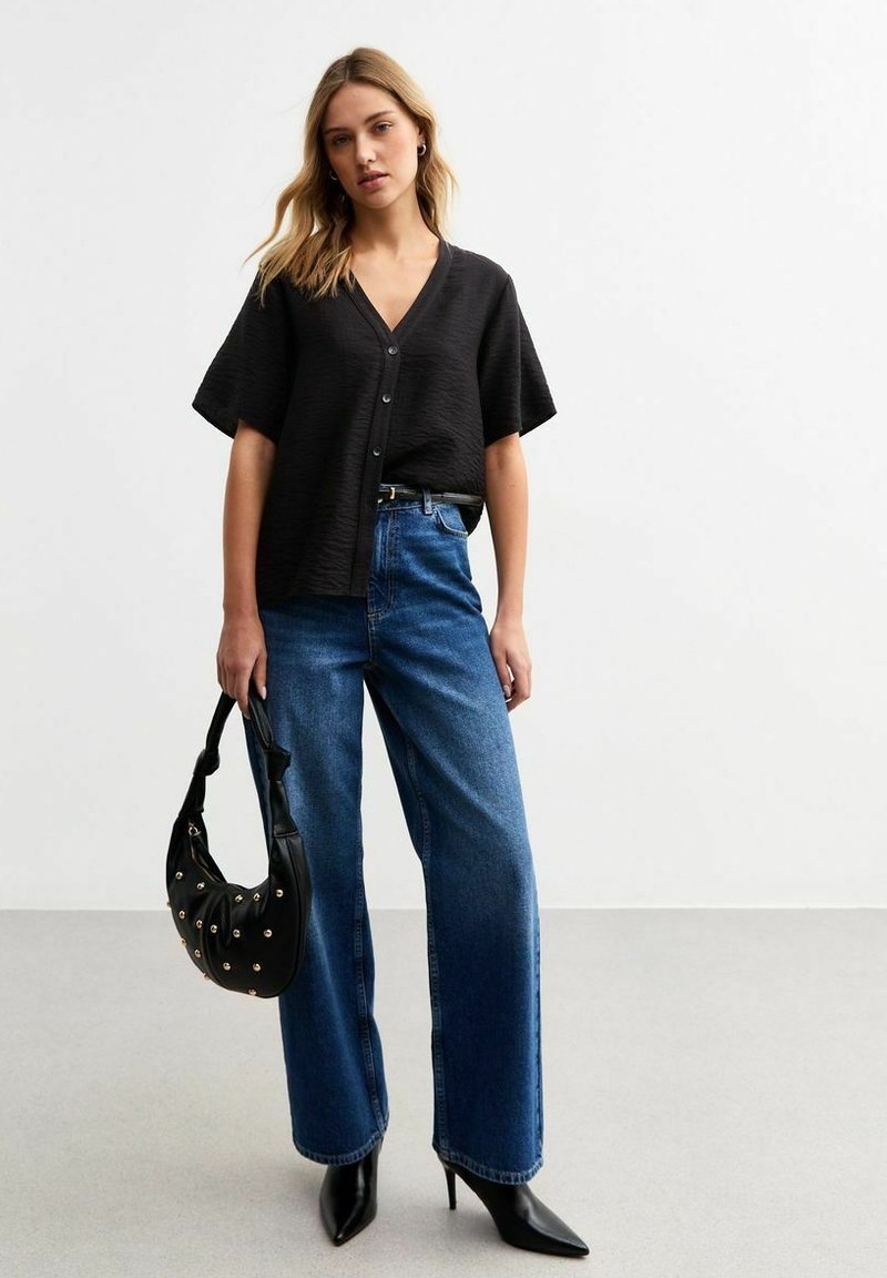 Zwarte, gestructureerde blouse met korte mouwen en een v-hals, met knoopsluiting, gecombineerd met hoge taille blauwe denim wijde jeans en puntige zwarte hakken.