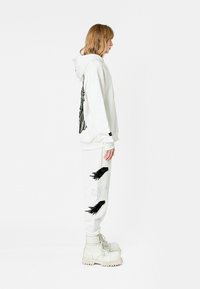 Sudadera blanca con bordado de alas negras en la parte posterior, combinada con pantalones de chándal blancos que presentan diseños de aves en negro y plata; botas blancas gruesas.