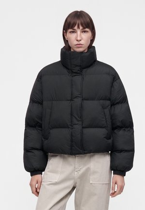 Frau mit glatten braunen Haaren trägt eine schwarze Steppjacke und hellbeige Hose, steht vor einem schlichten weißen Hintergrund.