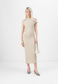 Beige ribba neulemekko yhdellä olkapäällä, tiukka muoto ja pehmeä rakenne. Mallilla on kädessään pieni valkoinen käsilaukku ja jalkineena teräväkärkiset korkokengät.