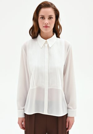 Giovane donna con capelli castani indossa una camicetta bianca a maniche lunghe con pieghe verticali e pantaloni marroni, in piedi davanti a uno sfondo bianco uniforme.