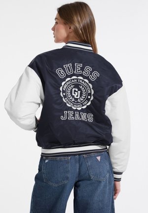 Laikinas ir baltas bomber tipo džemperis su "GUESS JEANS" logotipu ir apskritu emblema ant nugaros. Raibota apykaklė ir rankogaliai, blizgus paviršius, laisvas kirpimas.