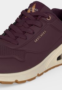 Gros plan d'une basket Skechers bordeaux avec semelle beige rembourrée, languette matelassée et détails de marque sur le côté et la languette.