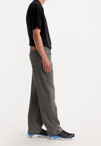 Jeans grigi a gamba larga in denim, con una toppa di pelle in vita, abbinati a una t-shirt nera semplice e scarpe sportive nere.