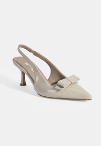 Talons aiguilles en cuir verni beige, avec un bout pointu, un talon de 7,5 cm et un accent de nœud décoratif sur le dessus. Fini lisse et forme structurée.