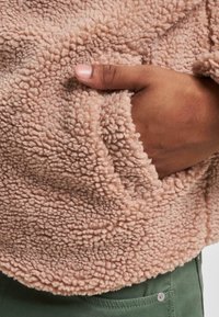 Polarowy sweter w jasnoróżowym kolorze o teksturowanej, puszystej powierzchni oraz z boczną kieszenią, zestawiony z zielonymi spodniami. Zbliżenie na materiał i kieszeń.