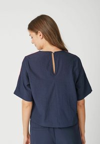 Donkerblauwe top met korte mouwen van een textuurstof, voorzien van een keyhole sluiting aan de achterkant en contrasterende accenten op de schouders.