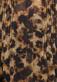 Camicia a maniche lunghe con motivo marrone e nero caratterizzata da pieghe, realizzata in tessuto leggero con una texture liscia. Quattro bottoni neri visibili.