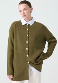 Cardigan in maglia a coste verde oliva con bottone frontale, caratterizzato da grandi bottoni crema, indossato sopra una camicia colletto a righe blu chiaro.