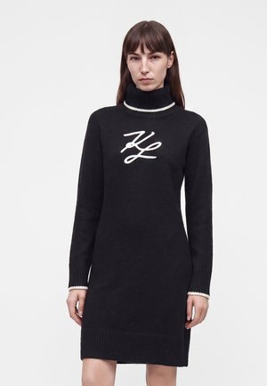 Žena s rovnými hnedými vlasmi, ktorá má na sebe čierne rolákové šaty so svetrovým materiálom s bielymi iniciálmi "KL" na hrudi a bielym lemom na golieri a manžetách.